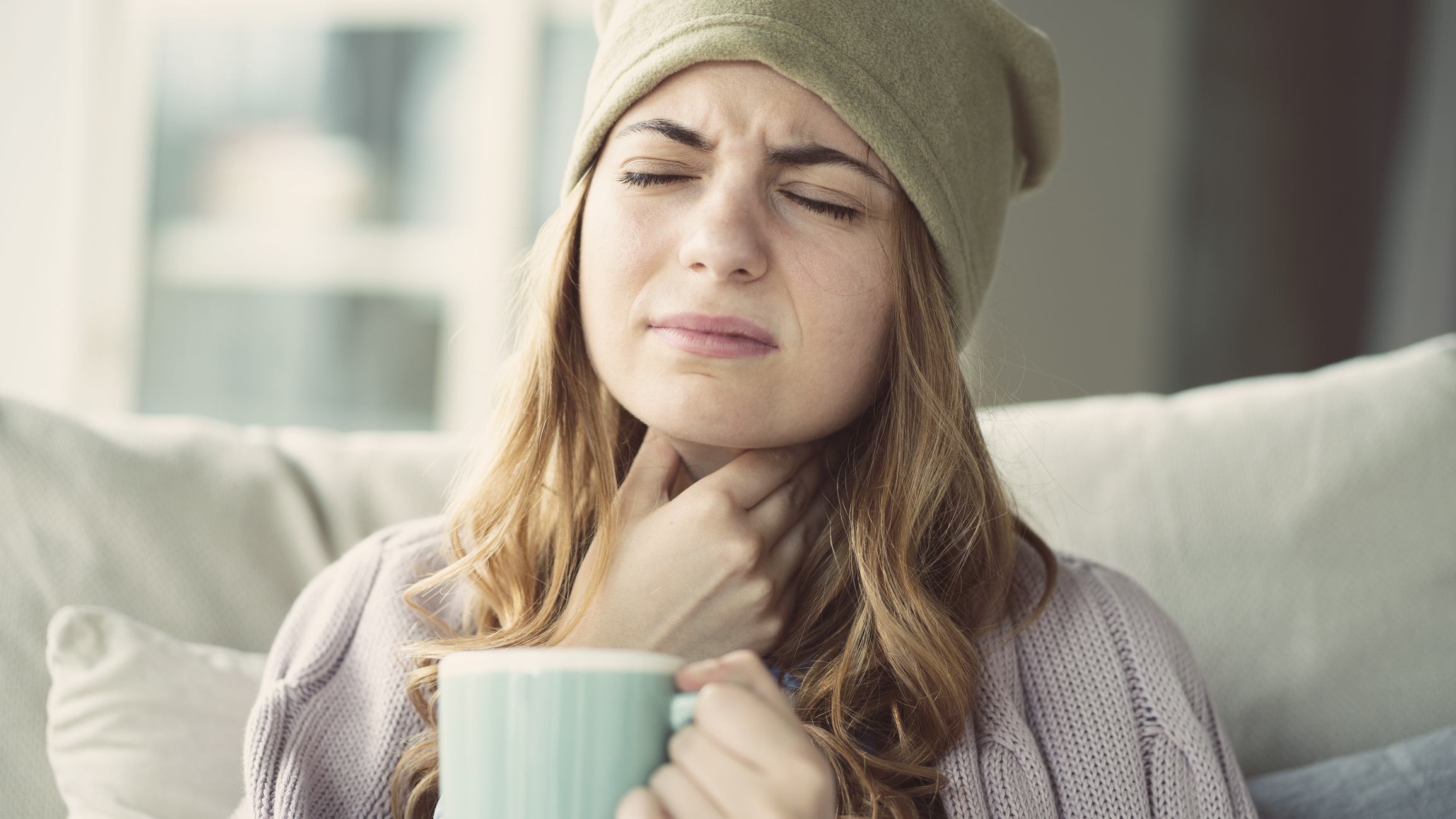 Woman in a hat & sitting on a couch is holding her sore throat with a mug in her other hand